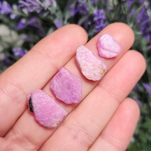 May include: Five pink rough cut gemstones, likely rubies, with a variety of shapes and sizes. The gemstones are displayed on a hand.