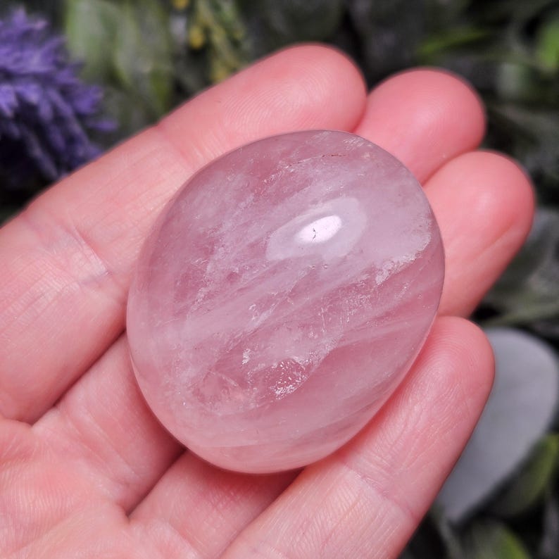 K&ouml;nnte beinhalten: Ein glatter, polierter, oval geformter Rosenquarz-Edelstein mit einem durchscheinenden, blassrosa Farbton. Der Stein weist nat&uuml;rliche Einschl&uuml;sse und eine gl&auml;nzende Oberfl&auml;che auf, die das Licht reflektiert und seine kristalline Struktur zeigt.