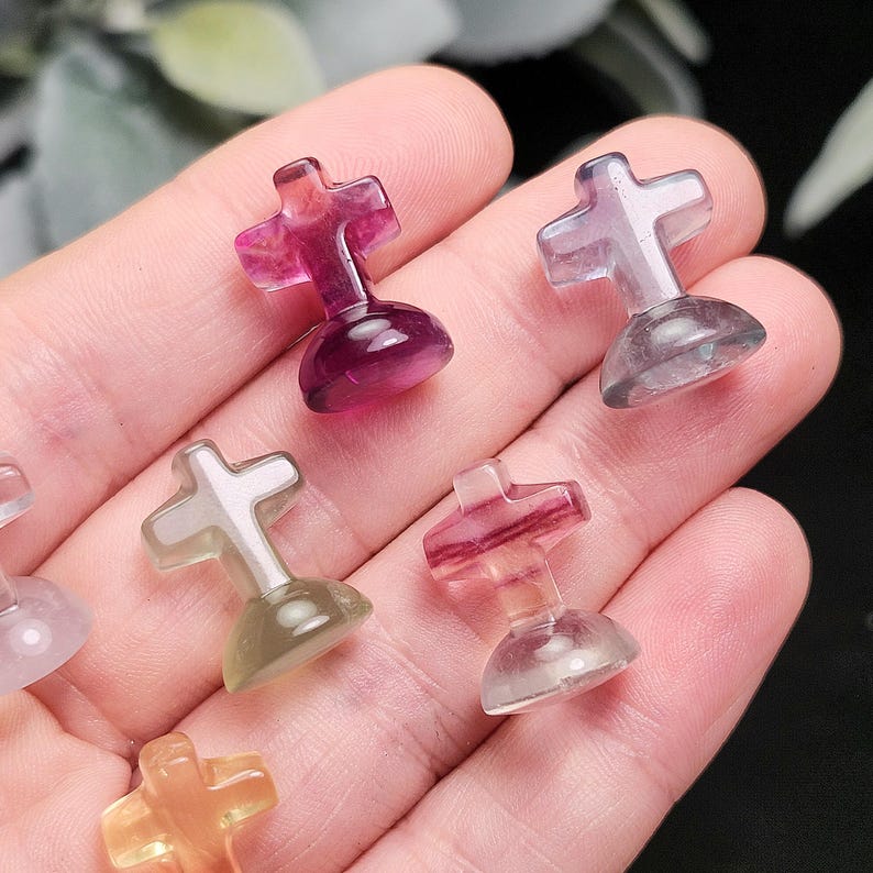 May include: A collection of small, translucent cross-shaped figurines in various colors, including purple, pink, and green. Each cross sits atop a rounded base. The figurines are held in a person's hand, showcasing their size and detail.