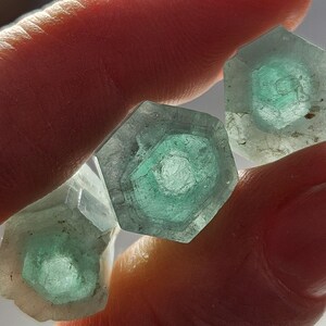 K&ouml;nnte beinhalten: Nahaufnahme von mehreren sechseckigen, durchscheinenden Kristallen. Jeder Kristall hat einen zentralen, mehrschichtigen, teal-farbenen Kern. Die Kristalle werden zwischen den Fingern gehalten, wobei das Licht durch sie scheint und ihre innere Struktur hervorhebt. Kristalle ca. 2-3 cm.