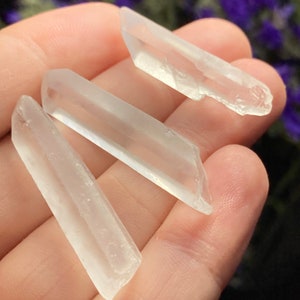 May include: Three clear quartz crystal points, also known as rock crystals, are displayed on a hand. The crystals are smooth and translucent.