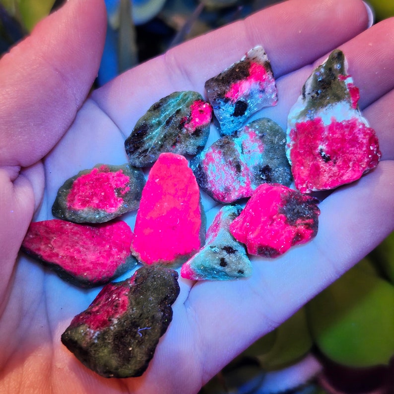 Pu&ograve; includere: Una collezione di pietre preziose grezze, non tagliate, di varie forme e dimensioni. Le pietre mostrano un mix di colori, tra cui rosa brillante, nero e verde, con alcune aree che brillano sotto una luce blu. Le pietre preziose sono tenute in una mano.