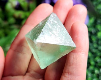 May include: A translucent, pale green fluorite crystal shaped as an octahedron with sharp, geometric edges and smooth, flat triangular faces. The mineral displays a light, minty green hue throughout its structure, showcasing natural clarity and internal crystalline patterns.