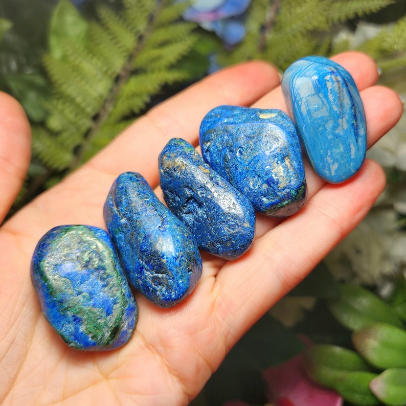 May include: A hand holds six polished azurite stones. The stones vary in shades of blue, with some displaying green and gold inclusions. The stones are smooth and rounded, showcasing natural textures and patterns. The background features greenery.