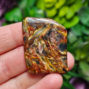 May include: A polished, rectangular-shaped pietersite gemstone cabochon featuring a swirling, chatoyant pattern of deep golden-yellow, dark brown, and black streaks with subtle hints of red, showcasing a smooth, reflective surface texture.