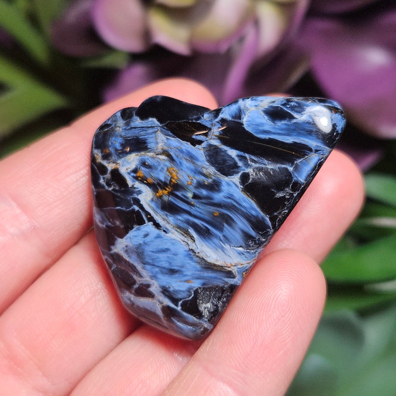 May include: A polished, heart-shaped pietersite gemstone featuring a swirling pattern of deep black and vibrant blue chatoyant fibers, accented with small, scattered flecks of golden-brown mineral inclusions across its smooth, reflective surface.