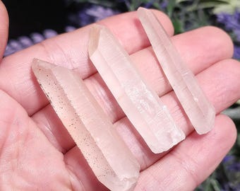 May include: Three clear quartz crystal points, each with a natural, rough texture. The crystals are a pale, translucent pink hue, with one showing small, dark inclusions. The crystals are held in a hand, with a blurred purple background.