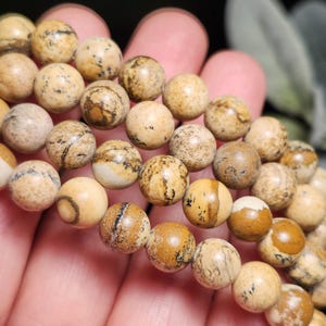 May include: Close-up of a strand of round, beige and brown jasper beads. The beads have a natural, earthy appearance with unique patterns and variations in color. The beads are strung together, suggesting a bracelet or necklace.