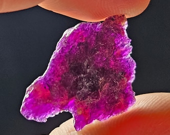 Pode incluir: Uma gema translúcida vibrante, de forma irregular, com um tom magenta profundo. As bordas são ásperas e irregulares, e a pedra é segurada entre dois dedos contra um fundo escuro. A cor da gema varia do roxo escuro ao rosa brilhante.