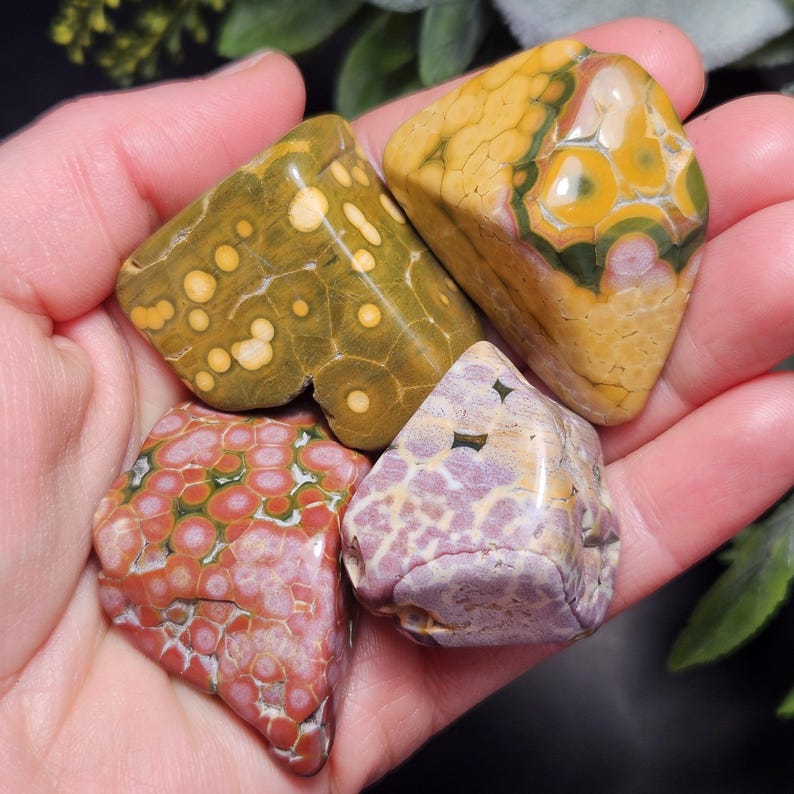 May include: Four polished gemstones in shades of green, yellow, pink, and purple. The stones have unique patterns, including spots and circular designs. The gemstones are held in a hand, showcasing their natural colors and textures.