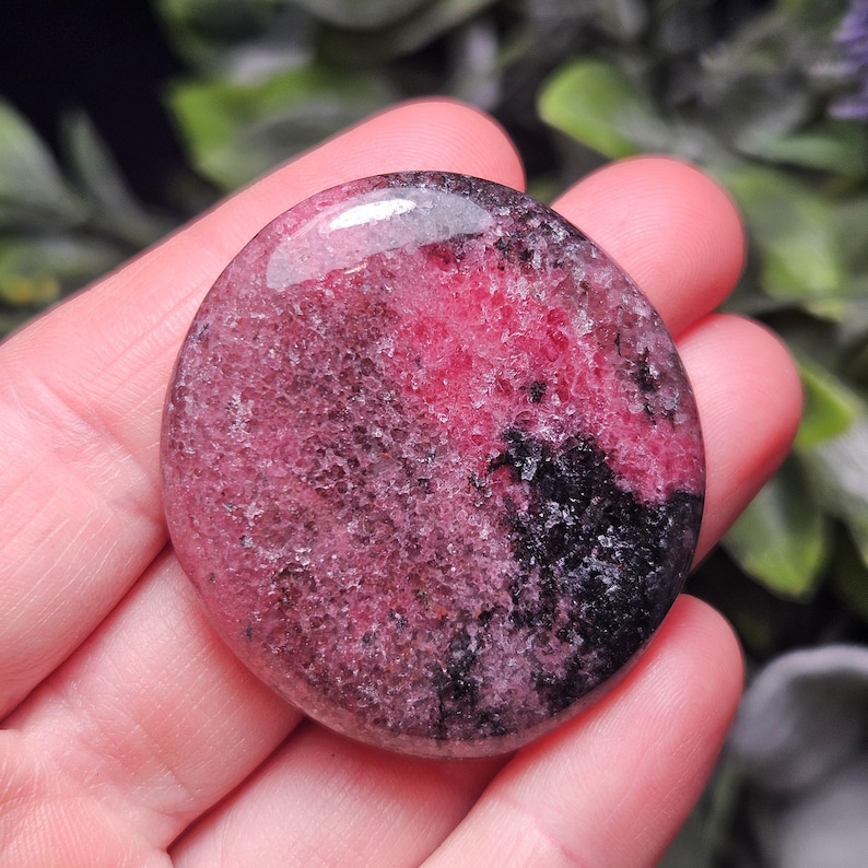 May include: A circular, polished rhodonite stone featuring a mottled pattern of vibrant pink, deep magenta, and dark black mineral inclusions. The surface has a smooth, glossy finish with a crystalline texture visible beneath the polished top layer.