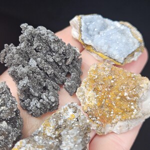 May include: A collection of raw mineral specimens, including a cluster of dark gray, crystalline formations and pieces with blue, yellow, and white hues. The minerals are displayed against a neutral background, showcasing their natural textures and colors.