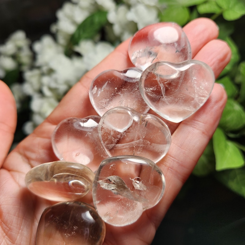 May include: A collection of clear quartz heart-shaped crystals, some with a slight golden hue, are displayed on a hand. The crystals are smooth and polished.