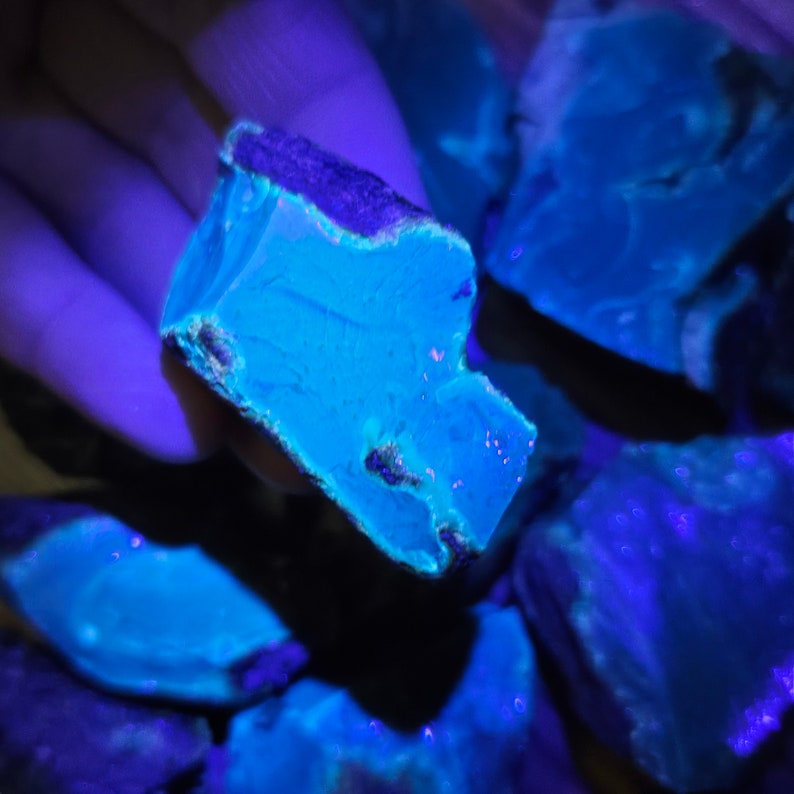 May include: Close-up of several blue-green mineral specimens under a UV light. The mineral glows a bright blue under the light.