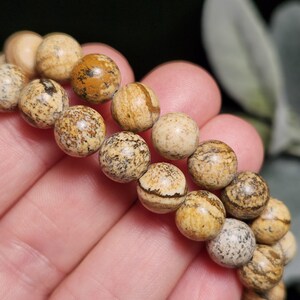 May include: A close-up of a beaded bracelet featuring round, natural stone beads. The beads are primarily tan and brown with unique patterns and textures. The bracelet is held in a person's hand, showcasing the details of the stone.