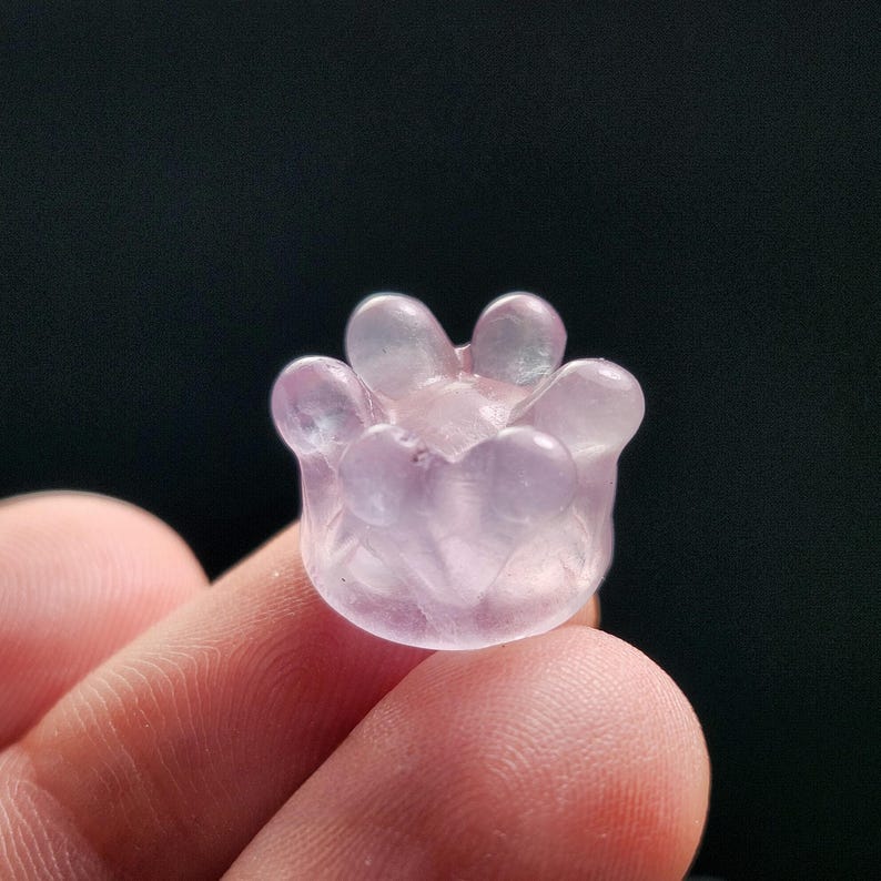 May include: A small, translucent, light purple flower-shaped carving. The carving has rounded petals and a detailed, textured surface. The object is held between fingers against a dark background, highlighting its delicate features.