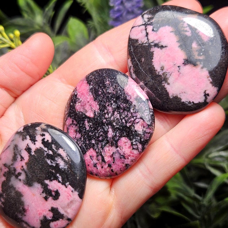 May include: Three polished, rounded rhodonite stones featuring a distinct pattern of vibrant pink and deep black mineral inclusions. Each stone has a smooth, glossy surface that reflects light, showcasing the natural contrast between the colors.