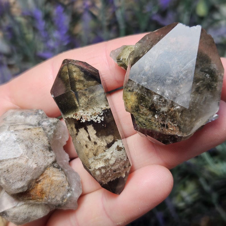 May include: A collection of raw, natural crystals in varying shapes and sizes. The crystals display a range of colors, including clear, brown, and green, with unique patterns and textures. The crystals are held in a hand, showcasing their natural beauty.