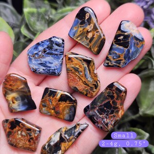 May include: A collection of polished pietersite stones featuring swirling patterns of deep blue, black, and golden brown. Each stone is small, weighing between 2-4g and measuring approximately 0.75 inches, displaying a smooth, reflective surface.