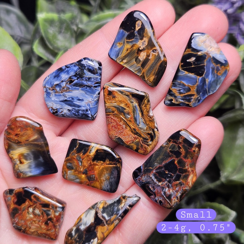 May include: A collection of polished pietersite stones featuring swirling patterns of deep blue, black, and golden brown. Each stone is small, weighing between 2-4g and measuring approximately 0.75 inches, displaying a smooth, reflective surface.