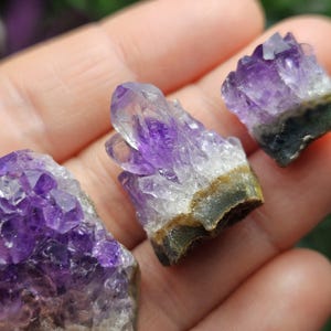 Pode incluir: Tr&ecirc;s pequenos aglomerados de cristais de ametista com um gradiente de cor roxo e claro. Os cristais s&atilde;o brutos e t&ecirc;m uma textura natural e &aacute;spera.