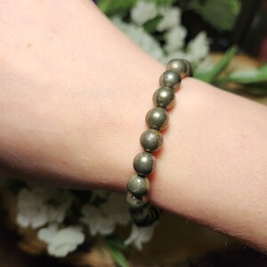 May include: A pyrite beaded bracelet. The bracelet features round, metallic gold-coloured beads strung together. The beads have a natural, textured appearance. The bracelet is shown on a person's wrist, with a soft background of white flowers and greenery.