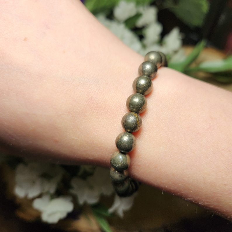 May include: A pyrite beaded bracelet. The bracelet features round, metallic gold-coloured beads strung together. The beads have a natural, textured appearance. The bracelet is shown on a person's wrist, with a soft background of white flowers and greenery.