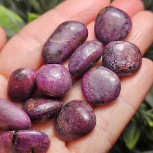 May include: A collection of polished, tumbled gemstones in shades of deep pink and purple. The stones vary in size and shape, with a smooth, glossy surface. Some stones have dark inclusions.
