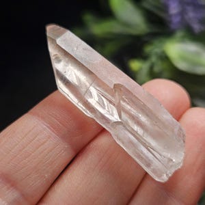 May include: A clear, elongated quartz crystal point with a pointed tip and natural faceted sides. The mineral specimen features internal inclusions and a transparent, glassy texture, showcasing a distinct geometric structure with sharp edges.