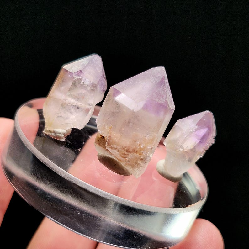 Puede incluir: Tres grupos de cristales de amatista sobre un soporte circular transparente. Los cristales son transl&uacute;cidos con puntas moradas y una base marr&oacute;n claro. El soporte est&aacute; sostenido por una mano sobre un fondo negro.