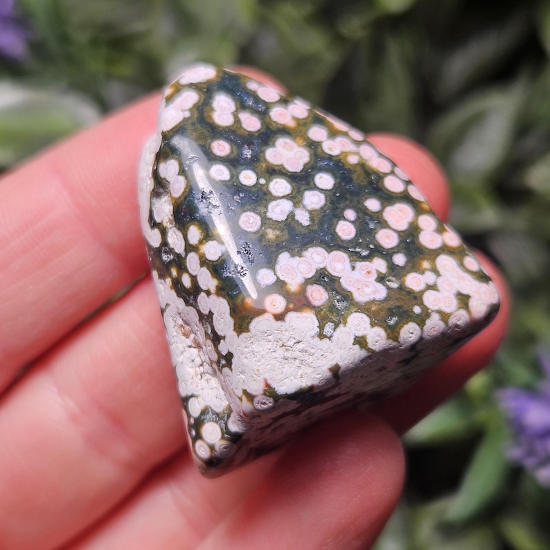 Puede incluir: Una piedra de jaspe oce&aacute;nico pulida que presenta una base de color verde oscuro con numerosos patrones circulares en tonos blancos y rosa suave. La piedra tiene una superficie lisa y brillante en la parte superior y una textura mate rugosa.