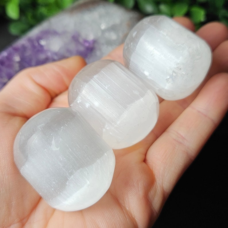 May include: Three white selenite tumbled stones, each with a smooth, rounded surface and a striated texture. The stones are arranged in a row, with the largest stone in the centre.