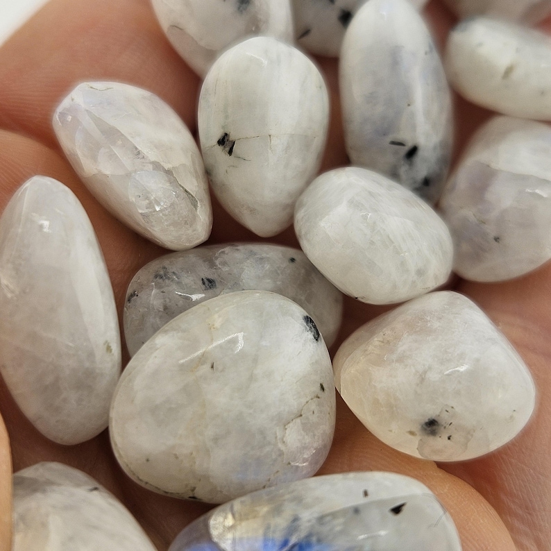 May include: A collection of polished moonstone gemstones. The stones are mainly white with subtle grey and black inclusions. The smooth, rounded shapes of the stones are visible, with a slight sheen. The gemstones are held in a hand.