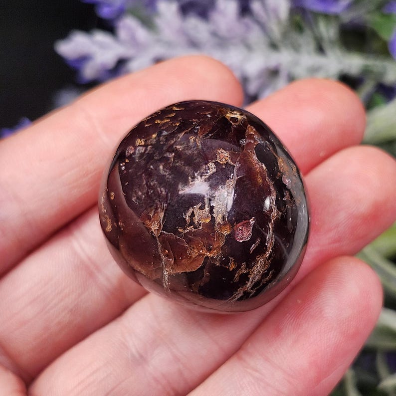 May include: A polished, dark burgundy gemstone with a rounded shape. The stone has a glossy surface with lighter brown and gold inclusions, held in an open hand. The background features blurred purple flowers.