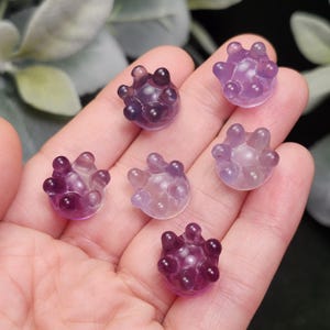 May include: Six small, translucent, purple-colored crystal spheres with rounded protrusions are displayed in a hand. The spheres vary in shades of purple, from light lavender to deep violet. The background is blurred, with hints of green foliage.