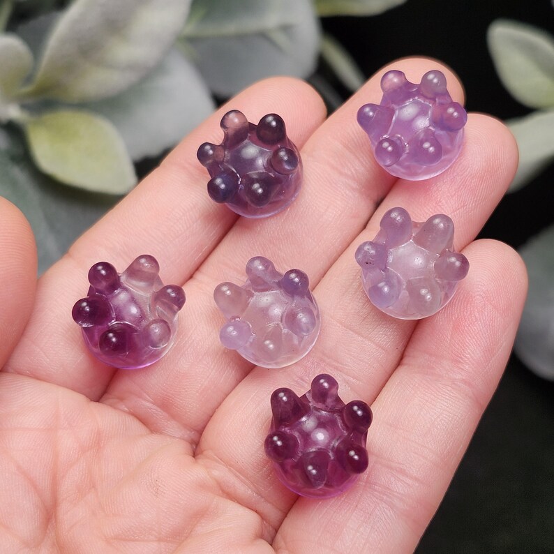 May include: Six small, translucent, purple-colored crystal spheres with rounded protrusions are displayed in a hand. The spheres vary in shades of purple, from light lavender to deep violet. The background is blurred, with hints of green foliage.