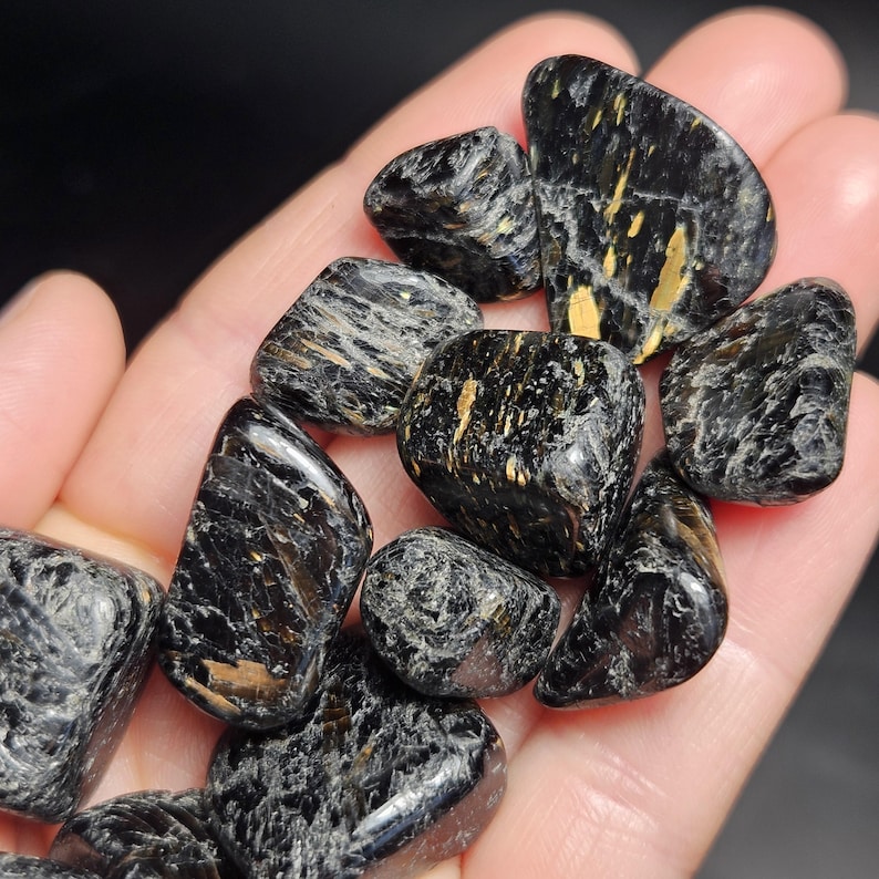 May include: A collection of polished black stones with gold-colored inclusions. The stones vary in size and shape, with smooth, rounded edges. The gemstones are held in a hand, showcasing their natural textures and colors. The overall aesthetic is earthy and organic.