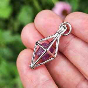 May include: A silver geometric cage pendant with a small, faceted, purple gemstone inside. The pendant is hanging from a silver chain.