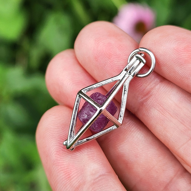 May include: A silver geometric cage pendant with a small, faceted, purple gemstone inside. The pendant is hanging from a silver chain.