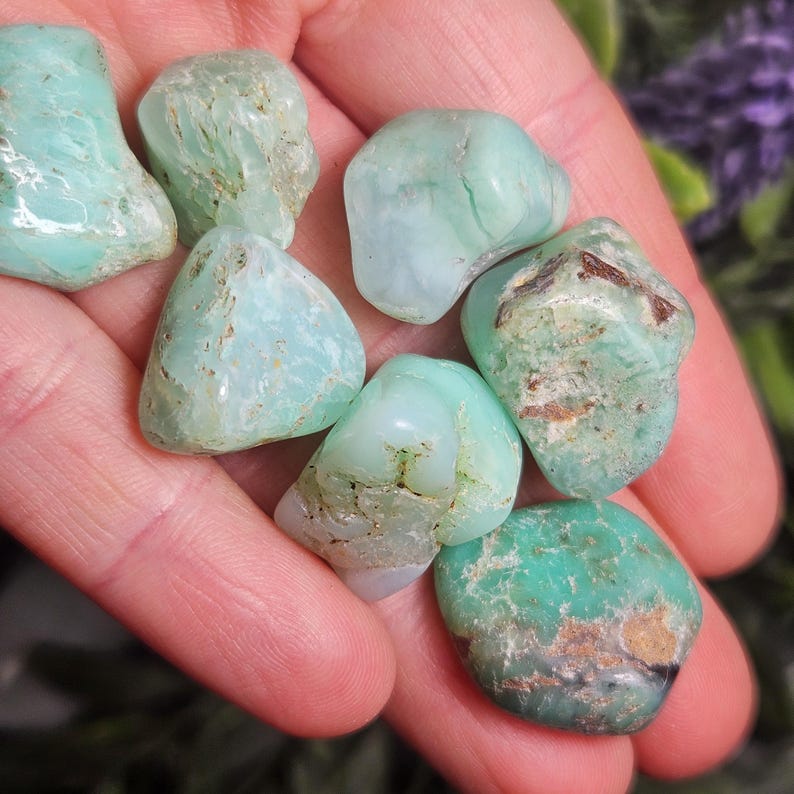 May include: A collection of tumbled chrysoprase stones featuring smooth, polished surfaces in various shades of mint green and seafoam green, with some stones displaying natural brown mineral inclusions and opaque, milky white patches.