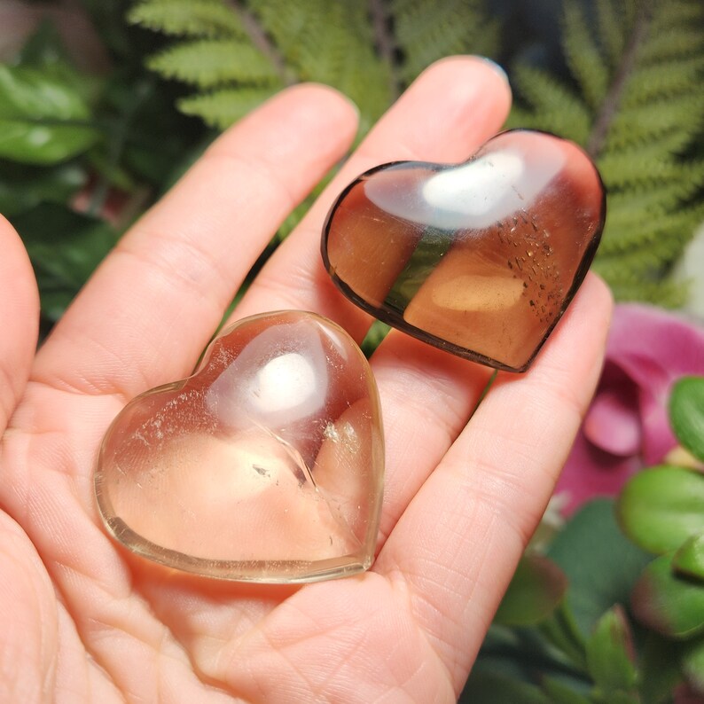 Puede incluir: Dos piedras de cristal en forma de coraz&oacute;n, una transparente y otra marr&oacute;n ahumada, se sostienen en una mano. Las piedras son lisas y pulidas.