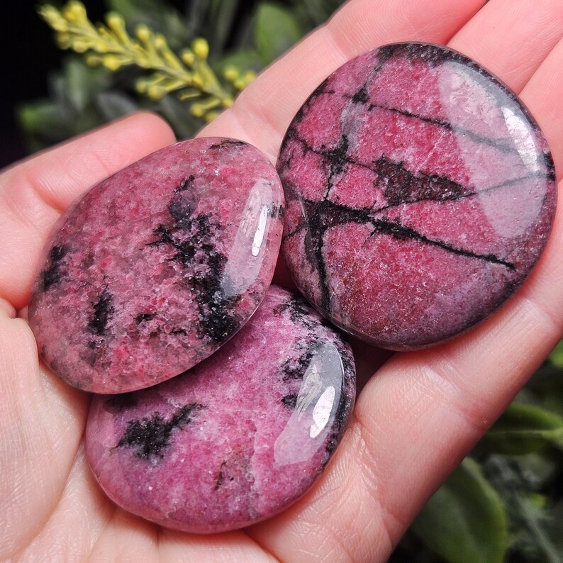 May include: Three polished, rounded rhodonite stones featuring a vibrant magenta pink base color with distinct, irregular black manganese oxide veins running throughout the surface of each stone, creating a natural, high-contrast pattern.
