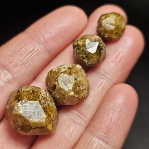May include: Four rough, golden-brown gemstones with faceted surfaces are displayed in a hand. The stones have a crystalline appearance and varying sizes. The background is dark, emphasising the gems' natural colour and texture.