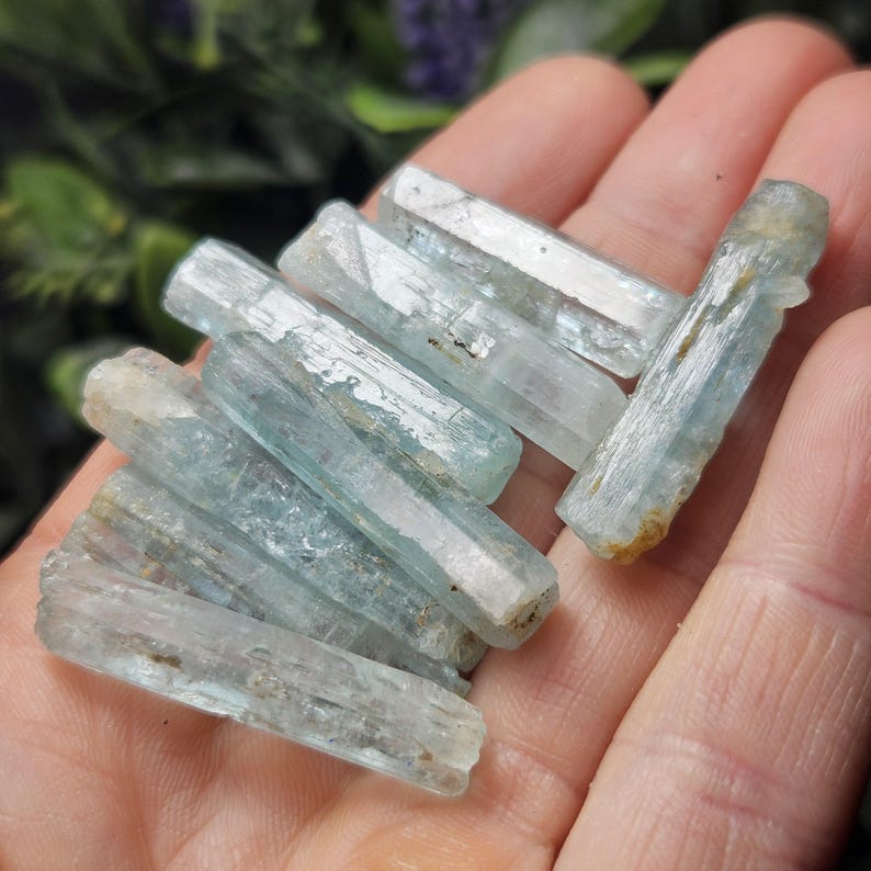 Puede incluir: Un conjunto de cristales de aguamarina en bruto, cada uno con un tono azul p&aacute;lido y una textura natural sin pulir. Los cristales se sostienen en una mano, mostrando sus formas prism&aacute;ticas alargadas y sutiles variaciones de color y claridad.