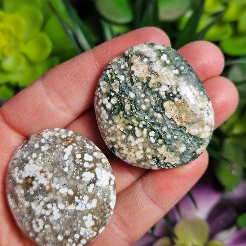 May include: Two polished ocean jasper stones. The stones have a mix of colors, including green, white, and tan. The ocean jasper has a speckled pattern and a smooth, rounded shape. The stones are held in a hand.