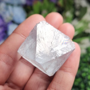 May include: A clear, iridescent, faceted crystal with a cubic shape. The crystal is held in a hand.
