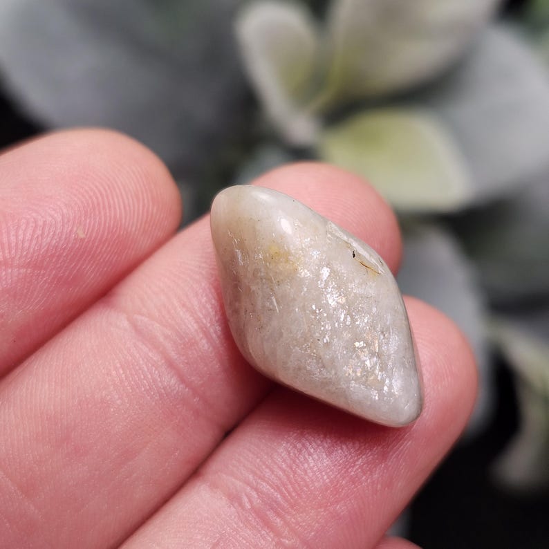 Puede incluir: Una gema oblonga pulida, de color blanco crema y amarillo p&aacute;lido. La piedra tiene una superficie lisa y reflectante y una peque&ntilde;a inclusi&oacute;n oscura. La gema se sostiene en la mano de una persona, con un fondo borroso verde y gris.