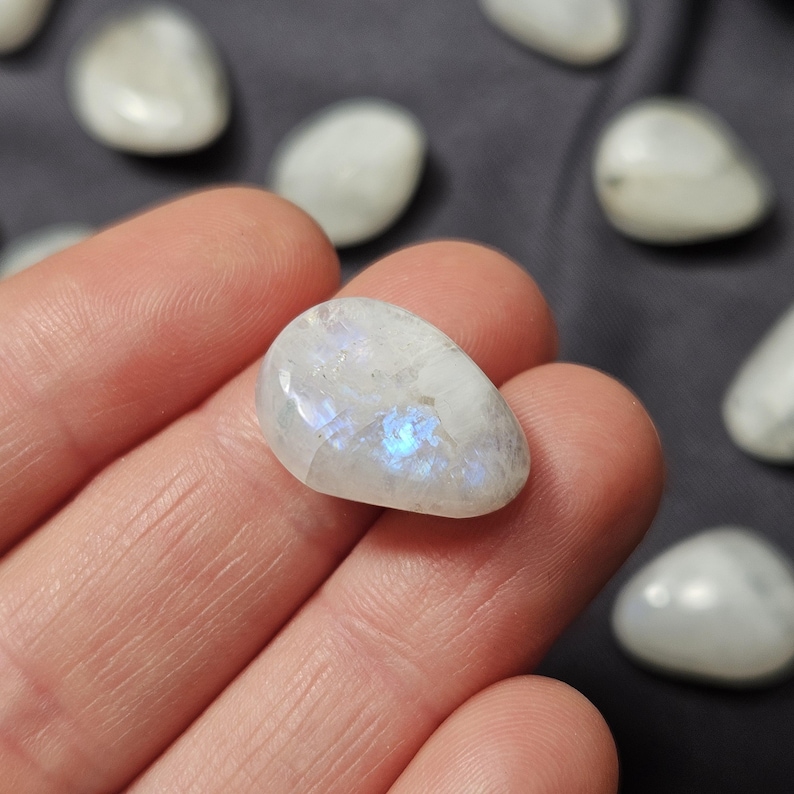 May include: A polished, oval moonstone displaying iridescent blue and white hues. The gemstone is held in a hand, with other moonstones in the background. The stones have a smooth, rounded form. The background is a dark grey.