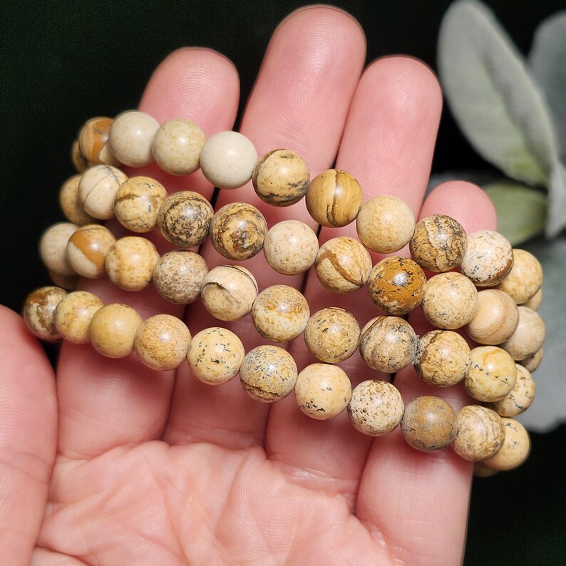 May include: Beaded bracelets featuring natural stone beads in shades of tan, brown, and cream. The round beads have unique patterns and textures, strung together to create multiple strands. The bracelets are held in a hand, showcasing the earthy tones and natural aesthetic.