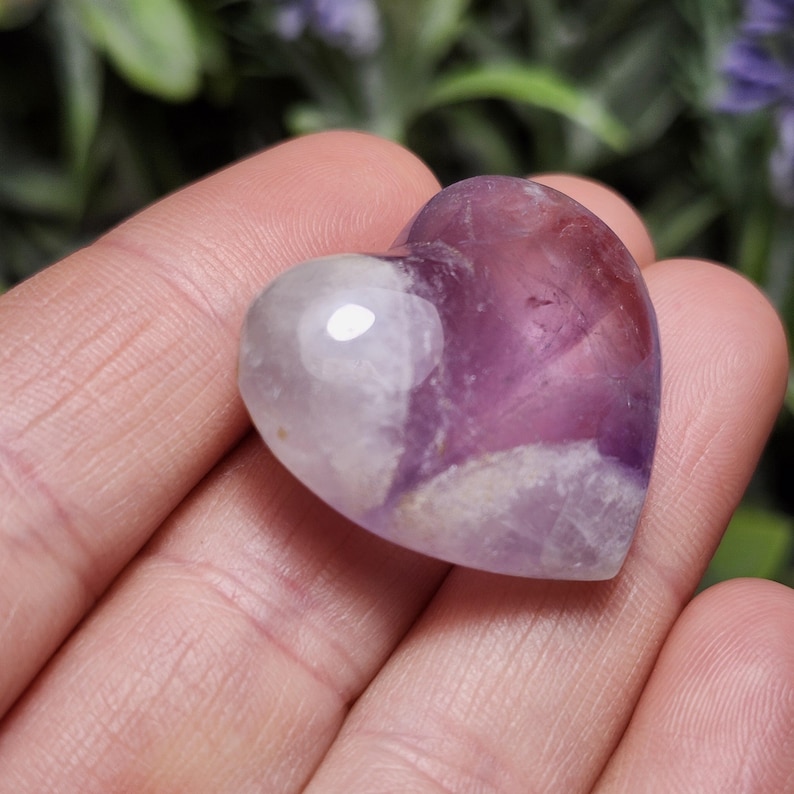 Puede incluir: Un cristal de amatista en forma de coraz&oacute;n con un patr&oacute;n de color p&uacute;rpura y blanco. El cristal es liso y pulido.
